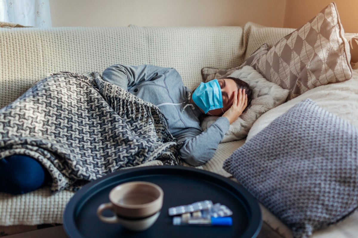 Frau liegt mit Maske krank auf dem Sofa.