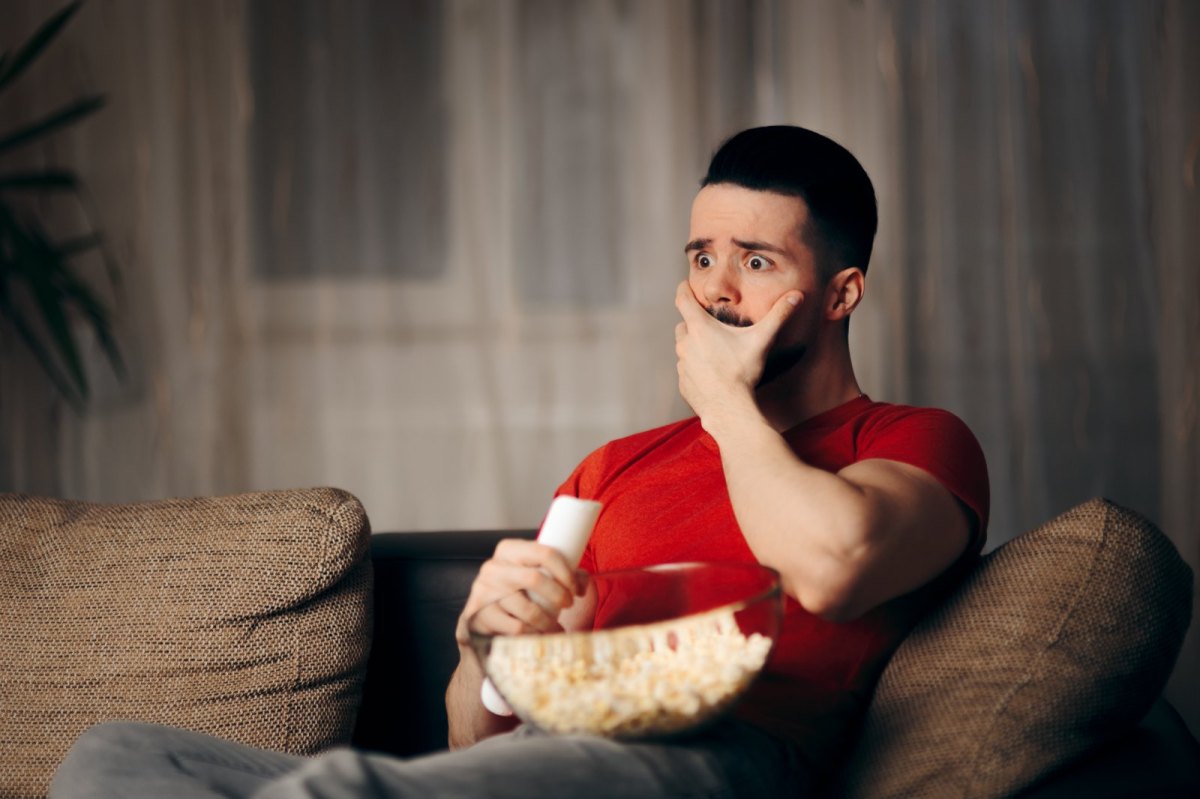 Man schaut einen Film und sitzt dabei auf dem Sofa. Er hat eine Schüssel Popcorn auf dem Schoß und ist erschrocken.