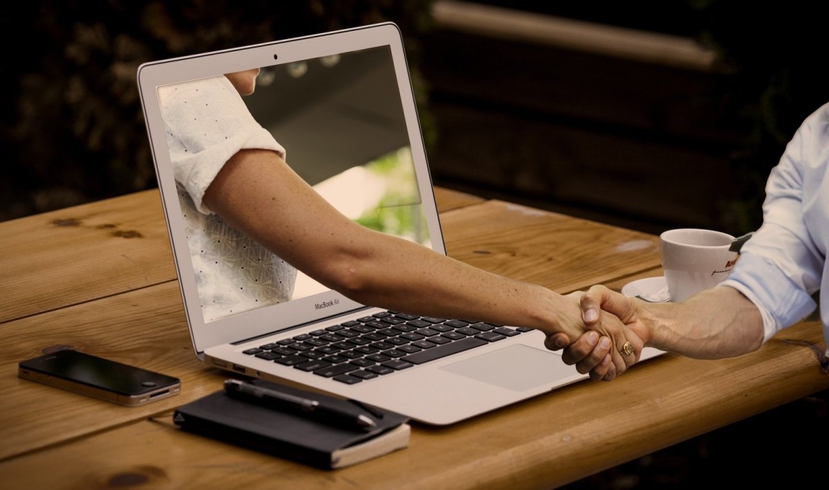 Eine Hand reicht einer anderen durch einen Computer hindurch die Hand.