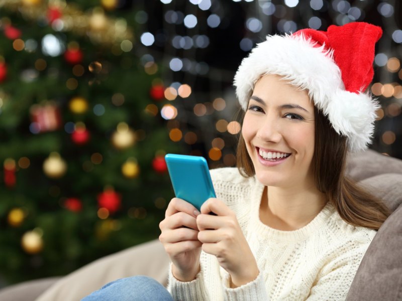 Frau sitzt mit ihrem Handy in einem Weihnachtssetting auf der Couch und guckt in die Kamera.