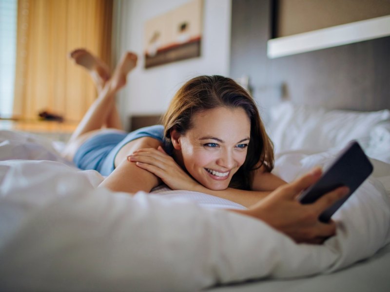 Frau liegt auf dem Bett in einem Hotelzimmer und hält dabei ein Handy in der Hand.