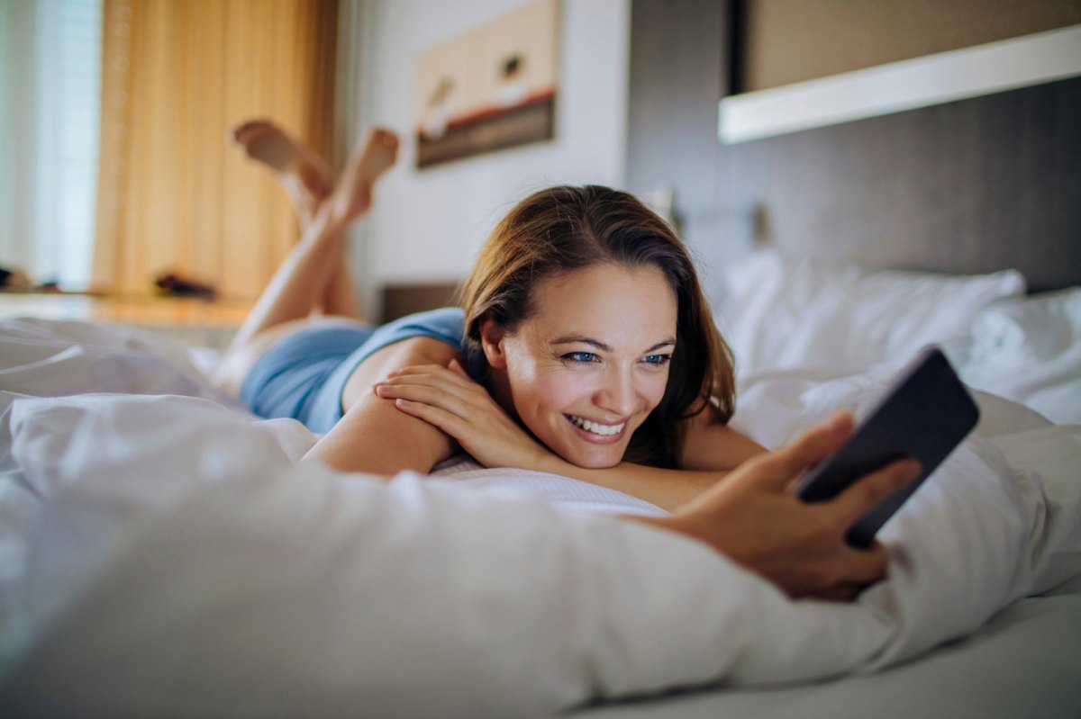 Frau liegt auf dem Bett in einem Hotelzimmer und hält dabei ein Handy in der Hand.