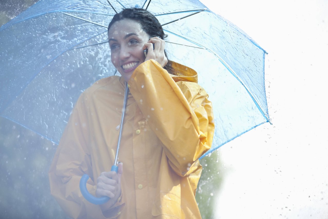 Frau mit Regenschirm und -jacke telefoniert mit ihrem Handy.