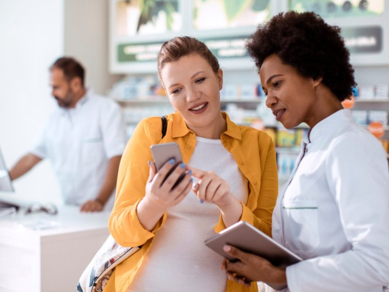 EIne Frau zeigt ihren Smartphone-Bildschirm einer Apothekerin.