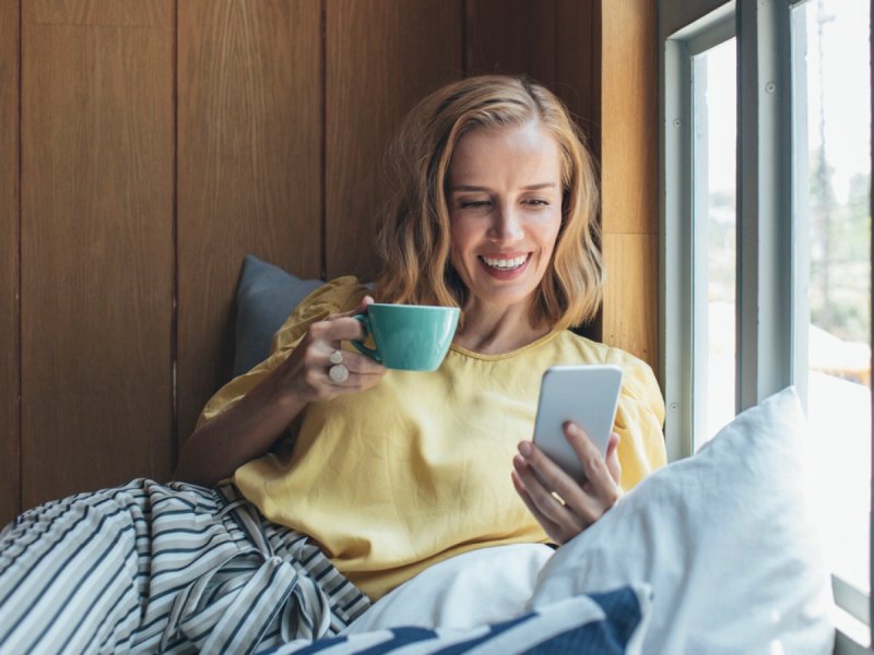 Frau sitzt mit Kaffee und einem Handy in der Hand gemütlich am Fenster.