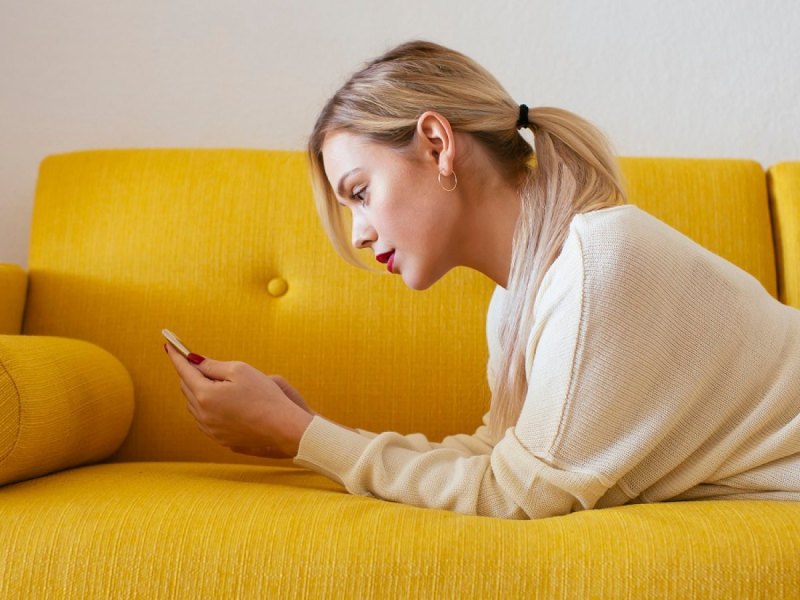 Frau mit Handy auf einem gelben Sofa.