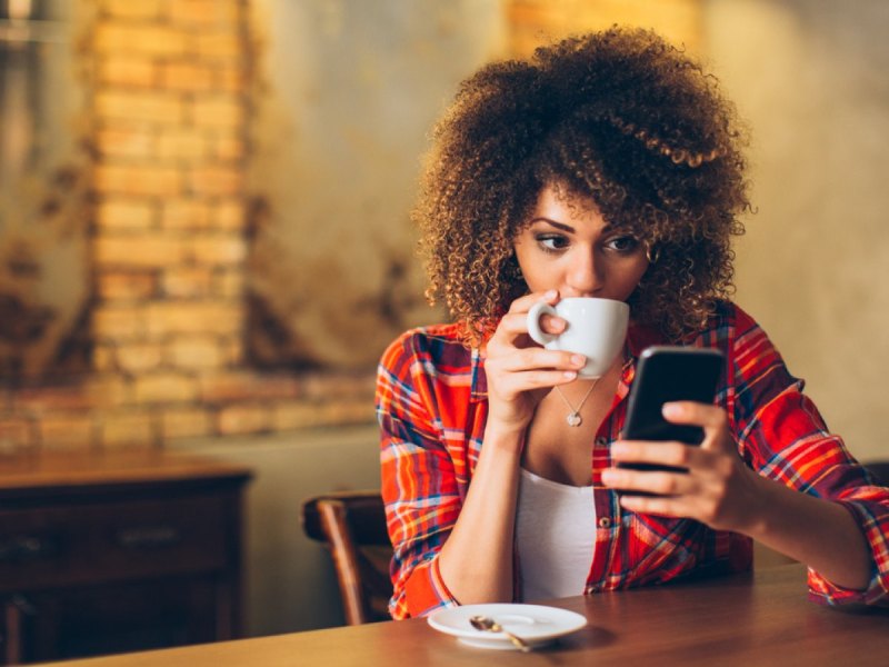 Frau mit Handy trinkt aus einer Tasse.