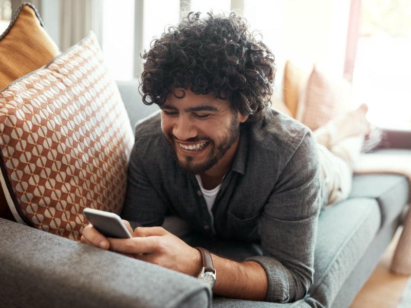 Mann liegt gemütlich auf dem Sofa und blickt glücklich auf sein Handy.