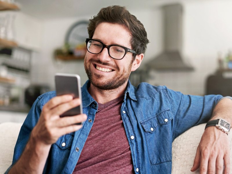 Mann sitzt auf dem Sofa und schaut lächelnd auf sein Handy.