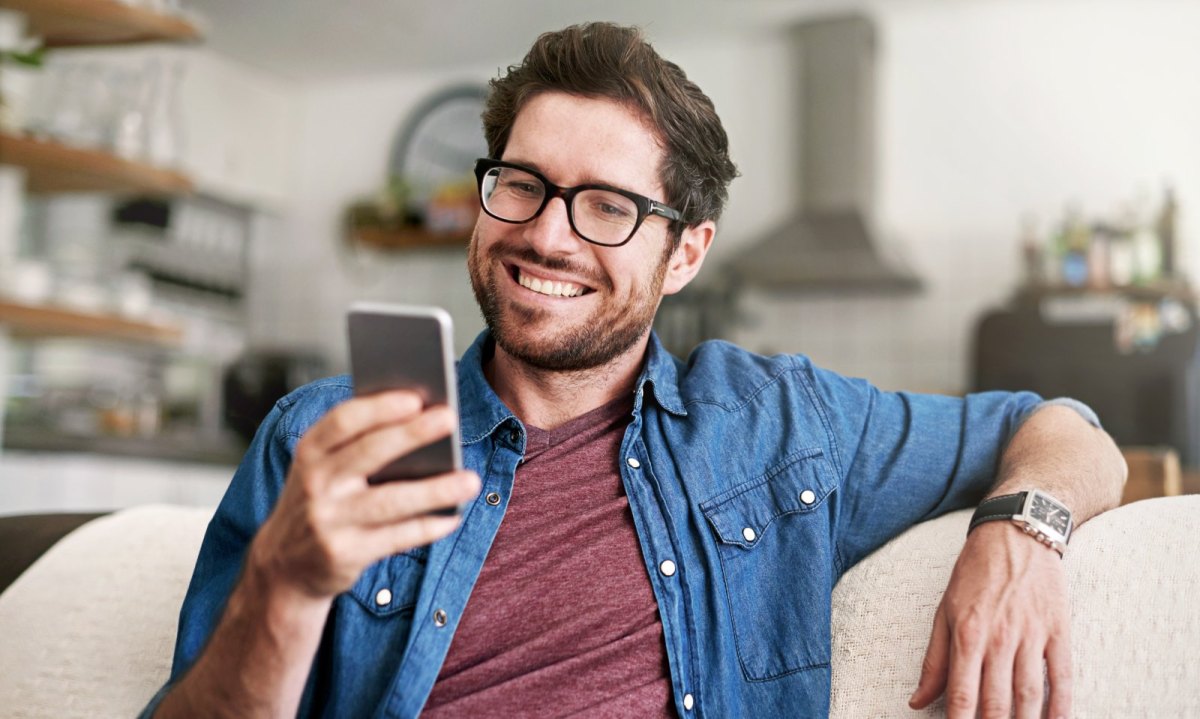 Mann sitzt auf dem Sofa und schaut lächelnd auf sein Handy.