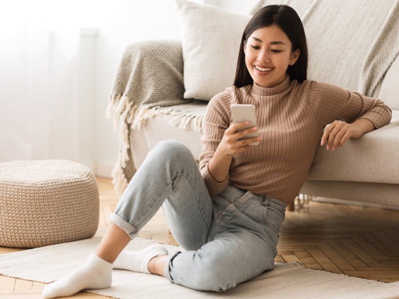 Frau lächelnd am Handy an eine Couch gelehnt.