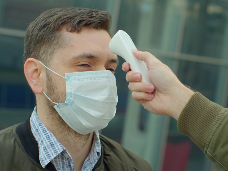 Mann mit Maske bekommt Fieberthermometer an die Stirn gehalten