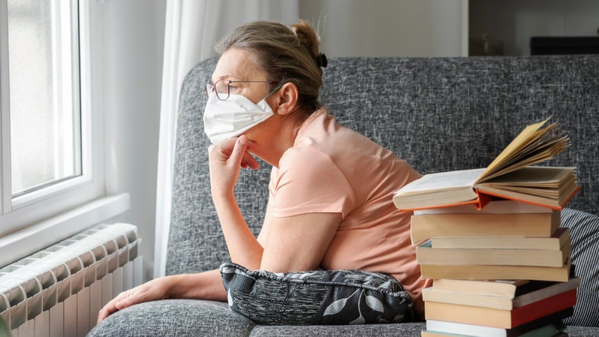 Frau blickt mit Mundschutz aus dem Fenster.