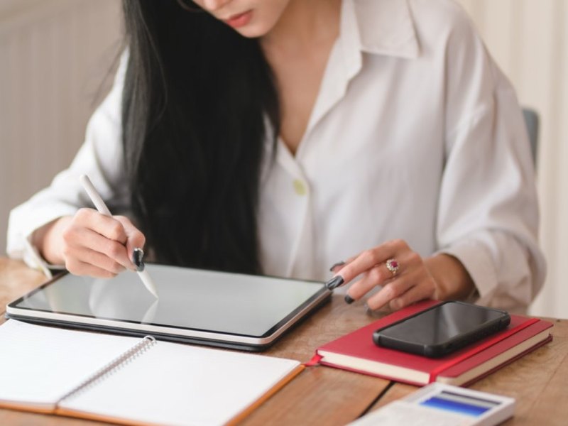 Eine Frau sitzt am Schreibtisch und schreibt mit einem Pen auf ihrem Tablet.