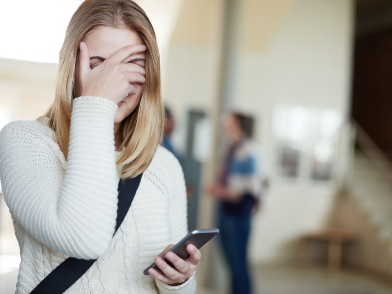 Eine Frau verdeckt peinlich berührt ihr Gesicht mit der Hand und hält das Handy in der anderen Hand.