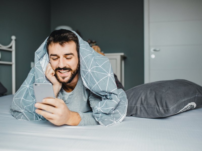 Mann liegt mit der Decke über den Schultern auf dem Bett und schaut fröhlich auf sein Handy.