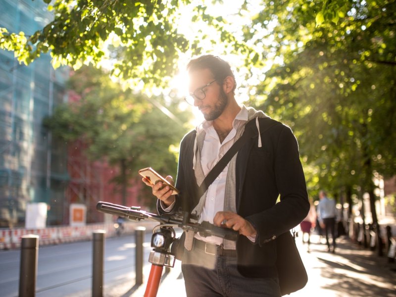 Mann blickt auf sein Handy und läuft währenddessen draußen mit seinem Fahrrad.