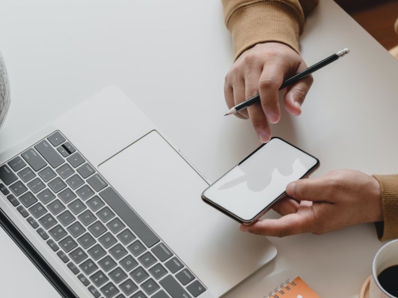 Ein Schreibtisch auf dem eine Person am Laptop und am Smartphone arbeitet.