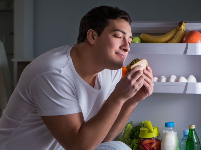 Mann isst Sandwich vor dem Kühlschrank.