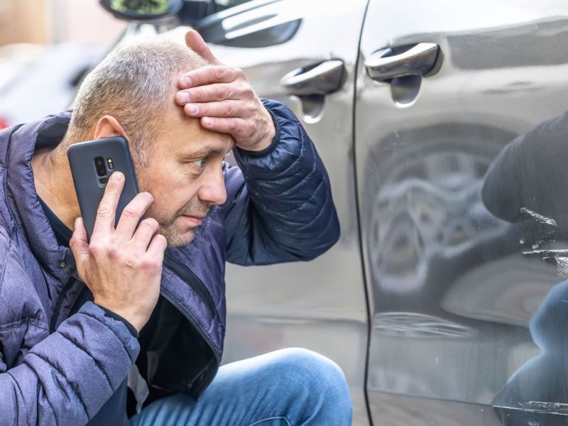 Mann telefoniert wÃ¤hrend er schockiert auf einen Kratzer an einem Auto schaut.