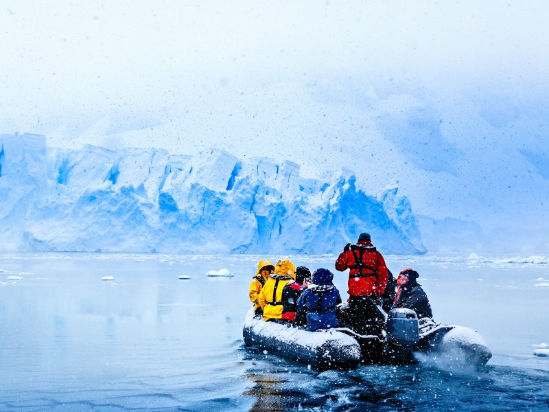 Schneefall über einem Boot mit Touristen, die auf die riesige blaue Gletscherwand im Hintergrund zufahren