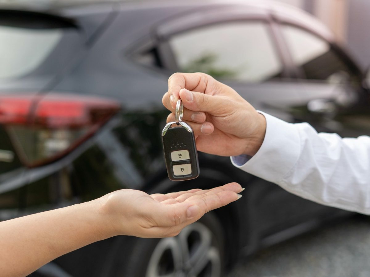 Person Ã¼bergibt einer anderen Person einen AutoschlÃ¼ssel. Im Hintergrund steht ein Auto.