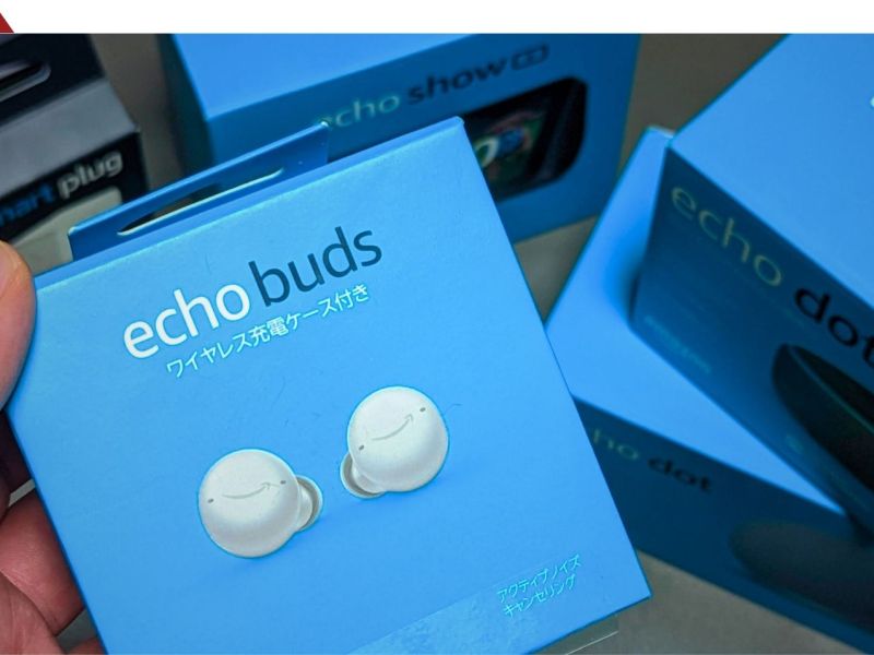 Eine Hand hÃ¤lt eine Packung Amazon Echo Buds. Dahinter liegen andere Produkte von Amazon.