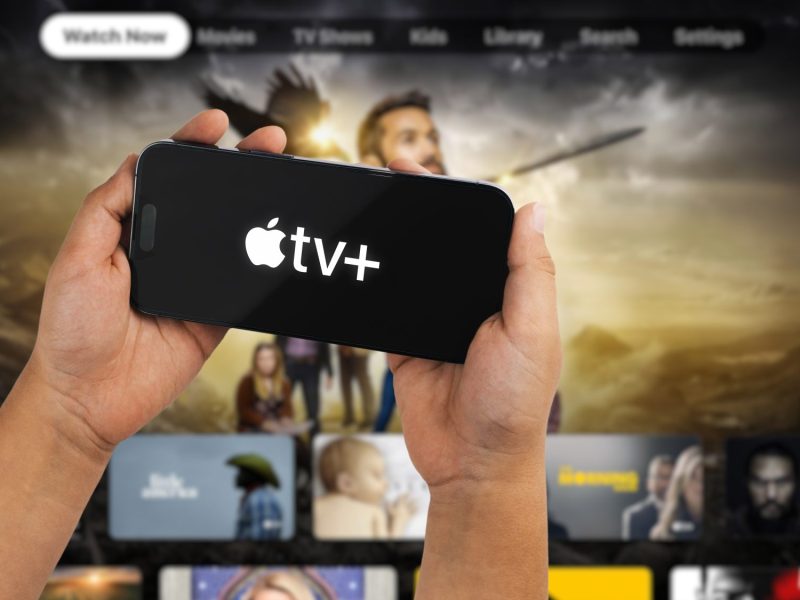 Auf einem Handydisplay wird das Apple TV+ Logo angezeigt. Im Hintergrund ist der Streaming-Dienst auf einem Fernseher geöffnet.