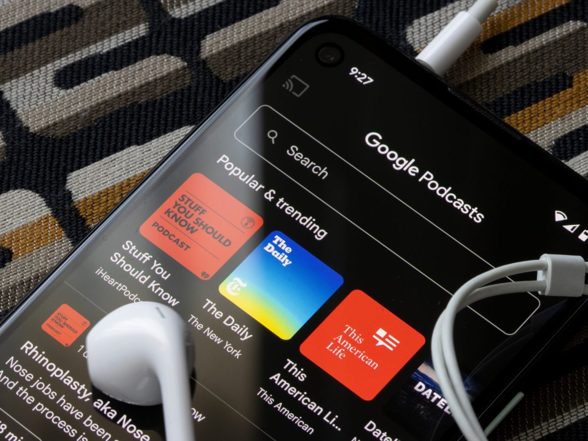 Auf einem Handy-Display ist die Google Podcasts-App geÃ¶ffnet. Auf dem Handy liegen KopfhÃ¶rer.