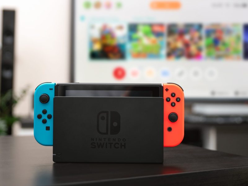 Eine Nintendo Switch-Konsole steht auf einem Tisch vor einem angeschalteten Fernseher.