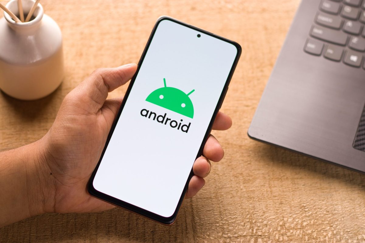 Eine Person hält ein Handy in der Hand. Auf dem Display ist das Android-Logo zu sehen. Im Hintergrund befindet sich ein Laptop auf einem Tisch.