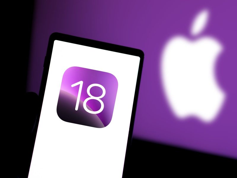 Handy zeigt iOS-18-Logo, im Hintergrund ist ein Apple-Logo zu sehen.
