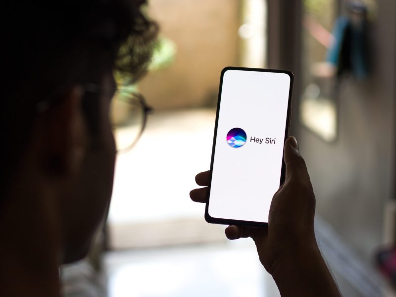 Eine Person hÃ¤lt ein Handy in der Hand, auf dessen Display Hey Siri angezeigt wird.