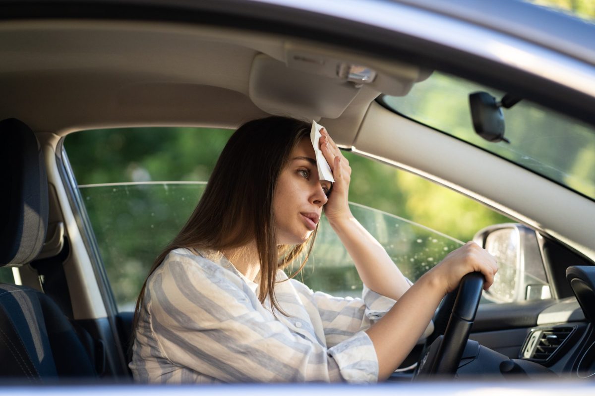 Eine Frau sitzt im Auto und hÃ¤lt sich ein Tuch an den Kopf und stÃ¼tzt sich auf das Lenkrad. Das Autofenster ist leicht geÃ¶ffnet.