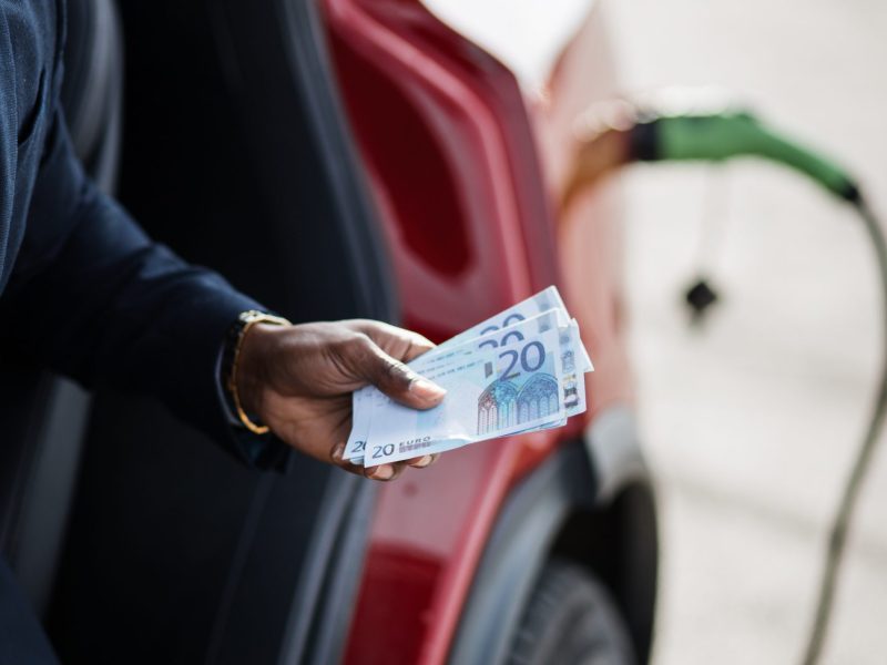 Person hÃ¤lt Geldscheine in der Hand. Im Hintergrund wird gerade ein Elektroauto geladen.