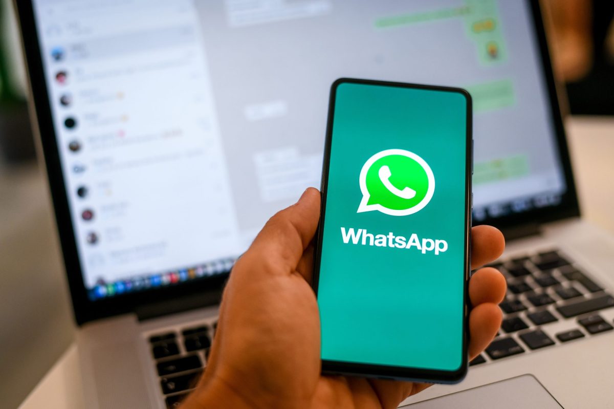 Person hÃ¤lt Handy in der Hand. Auf dem Bildschirm ist das WhatsApp-Logo zu sehen, im Hintergrund steht ein Laptop, auf dem WhatsApp geÃ¶ffnet ist.