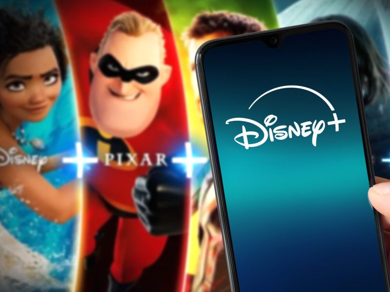 Auf einem Handy-Display wird das Disney+-Logo angezeigt. Im Hintergrund sind Charaktere aus Animationsfilmen zu sehen.