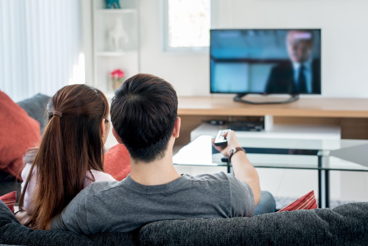 Zwei Personen sitzen auf einem Sofa und gucken auf einen Fernseher.