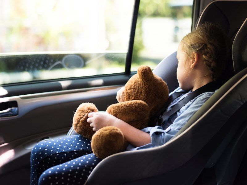 MÃ¤dchen sitzt in einem Auto-Kindersitz und hÃ¤lt einen Teddy in der Hand, wÃ¤hrend sie aus dem Fenster guckt.