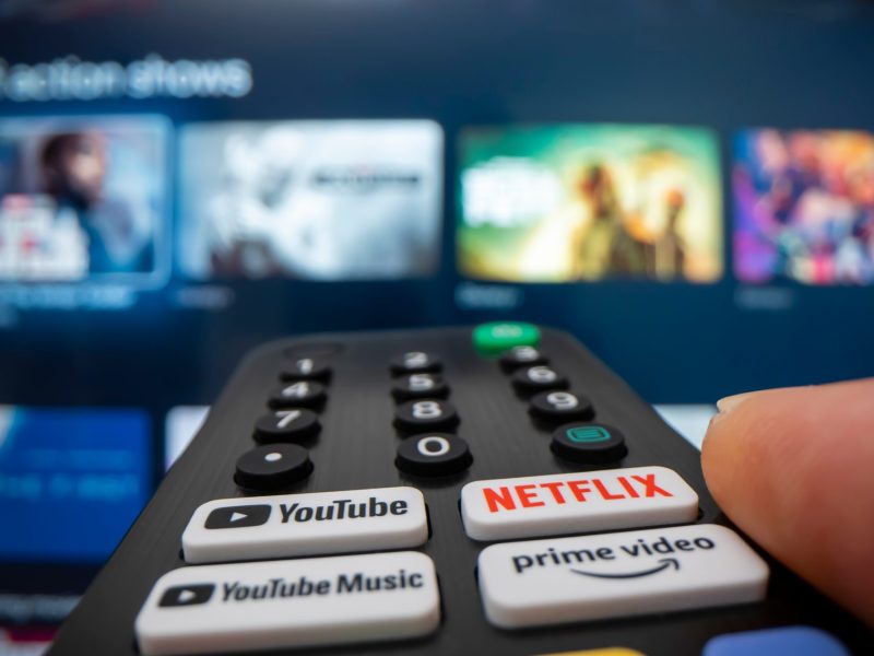 Person hält eine Fernbedienung auf einem Fernseher, auf dem verschiedene Streaming-Angebot angezeigt werden.