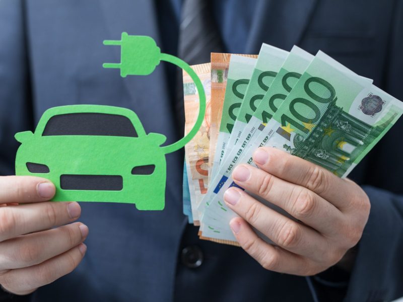 Eine Person hält in der einen Hand eine Zeichnung eines Elektroautos und in der anderen Hand mehrere Euroscheine.