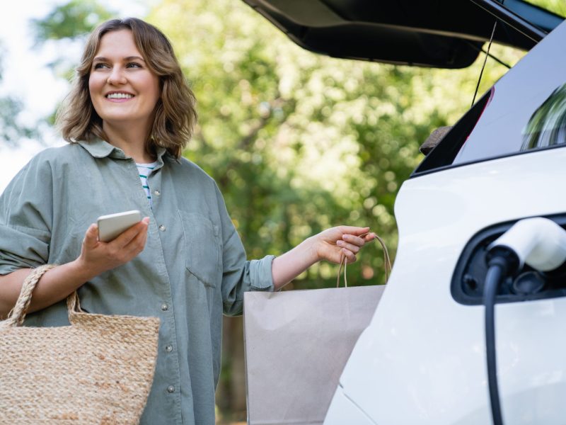 Frau lädt Einkäufe in den Kofferraum, während ihr Elektroauto geladen wird.