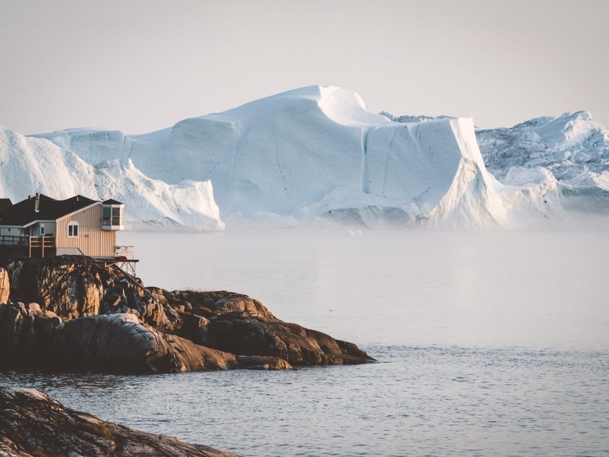 Eine Holzhütte steht an einer Felsküste. Im Hintergrund ist eine riesige Eisformation zu sehen.
