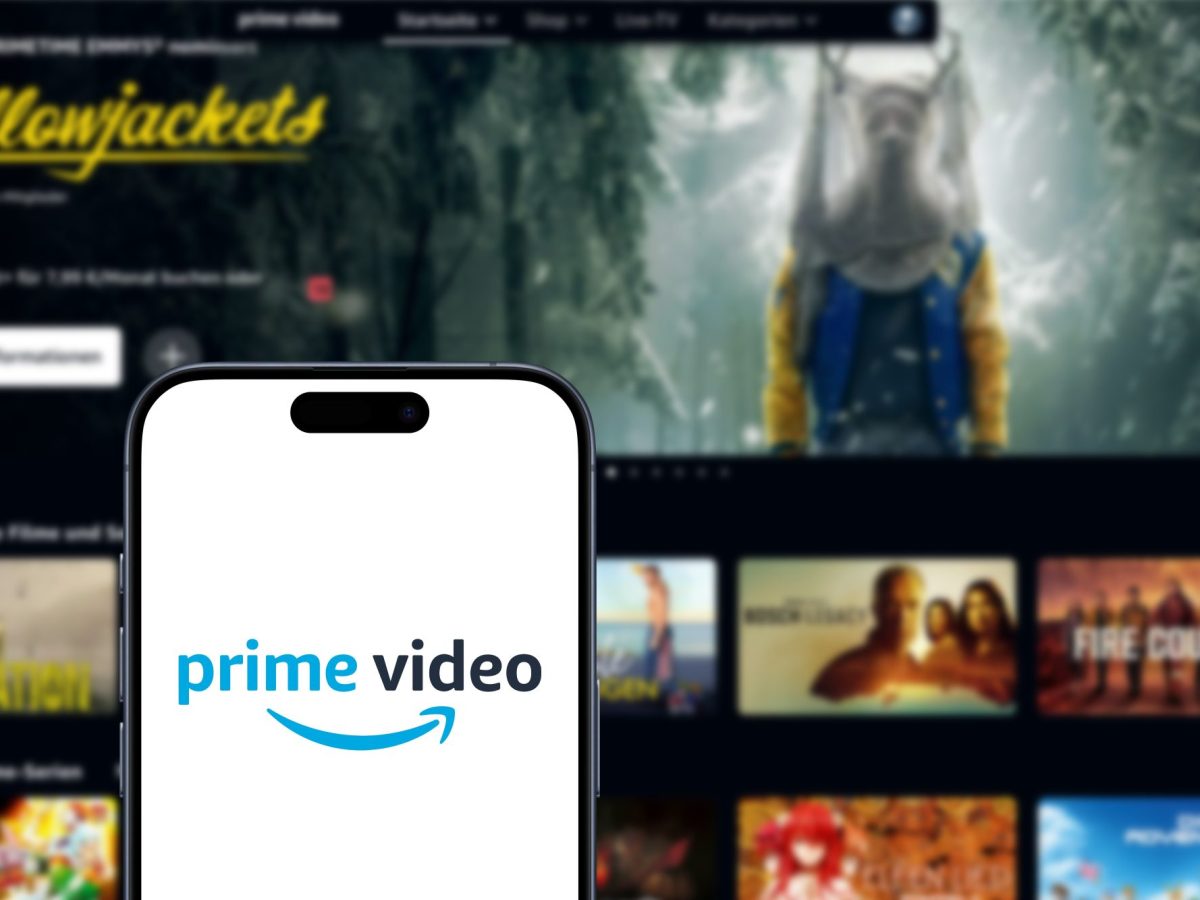 Auf einem Handy-Display wird das Amazon Prime Video-Logo angezeigt. Im Hintergrund ist ein Fernseher zu sehen, auf dem mehrere Video-Inhalte angezeigt.