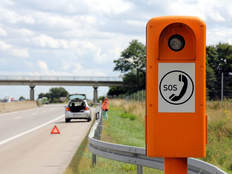 SOS-Säule an einer Autobahn. Im Hintergrund ist ein Auto zu sehen, das hinter einem Warndreieck auf dem Seitenstreifen steht.