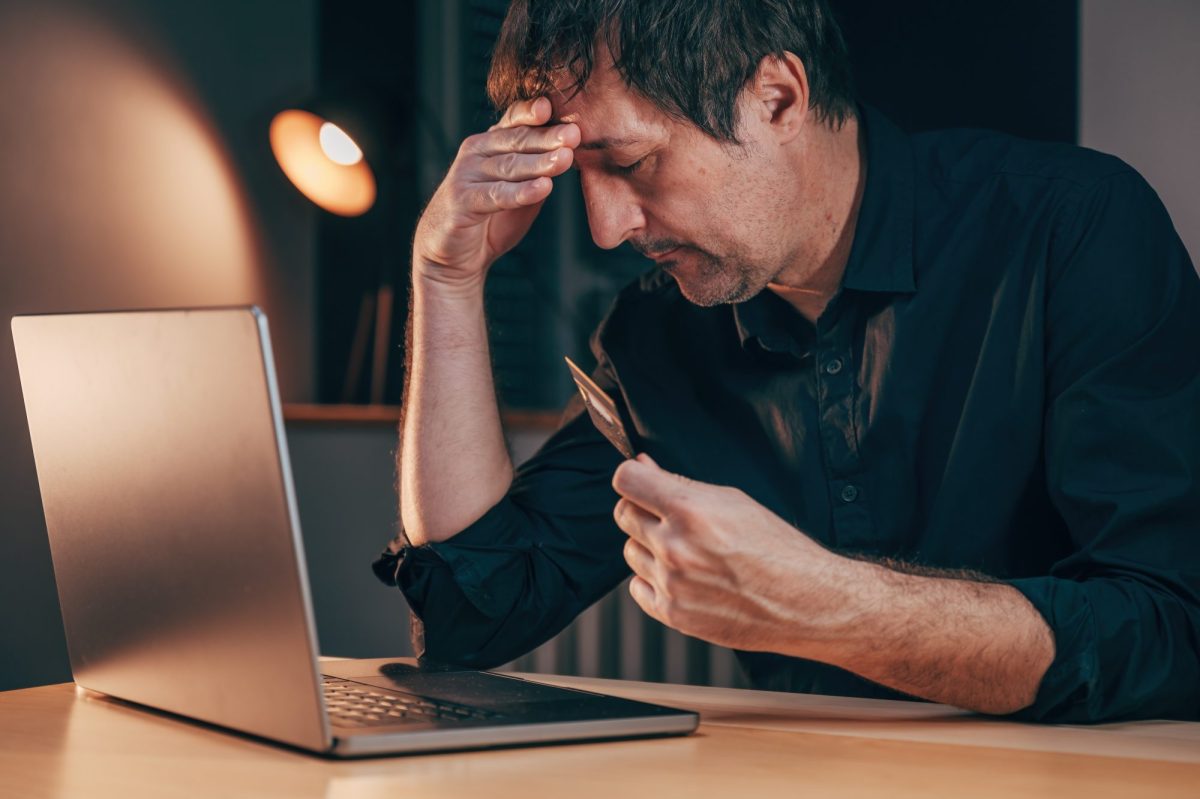 Mann hält verzweifelt die Hand an den Kopf, während er vor einem Laptop sitzt, mit einer Bankkarte in der Hand