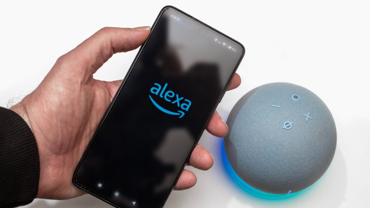 Person hält ein Handy, auf dessen Display das Alexa-Logo angezeigt wird. Daneben ist ein Amazon Echo Dot zu sehen.