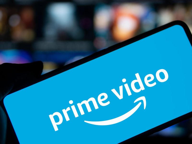 Amazon Prime Video-Logo wird auf einem Handy-Display angezeigt. Dahinter ist ein eingeschalteter Fernseher zu sehen.