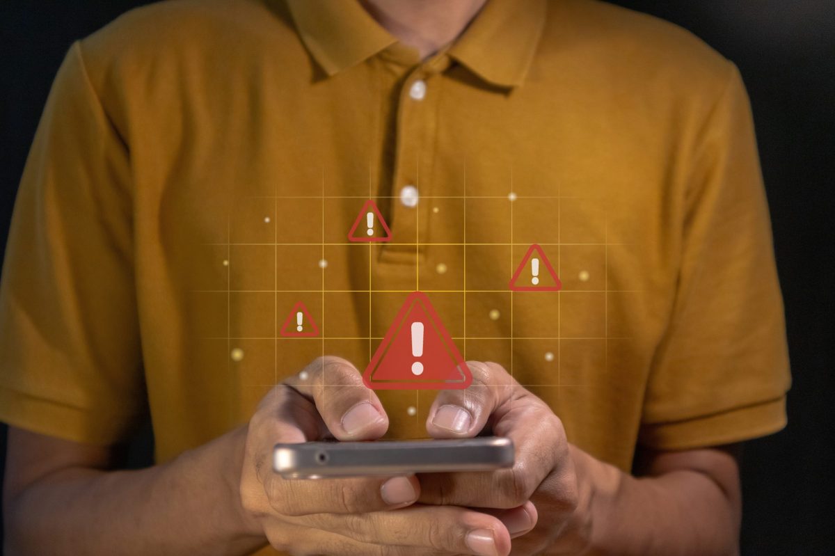 Person hält ein Handy in der Hand. Darüber ist ein rotes Ausrufezeichen zu sehen und symbolisiert Gefahr.