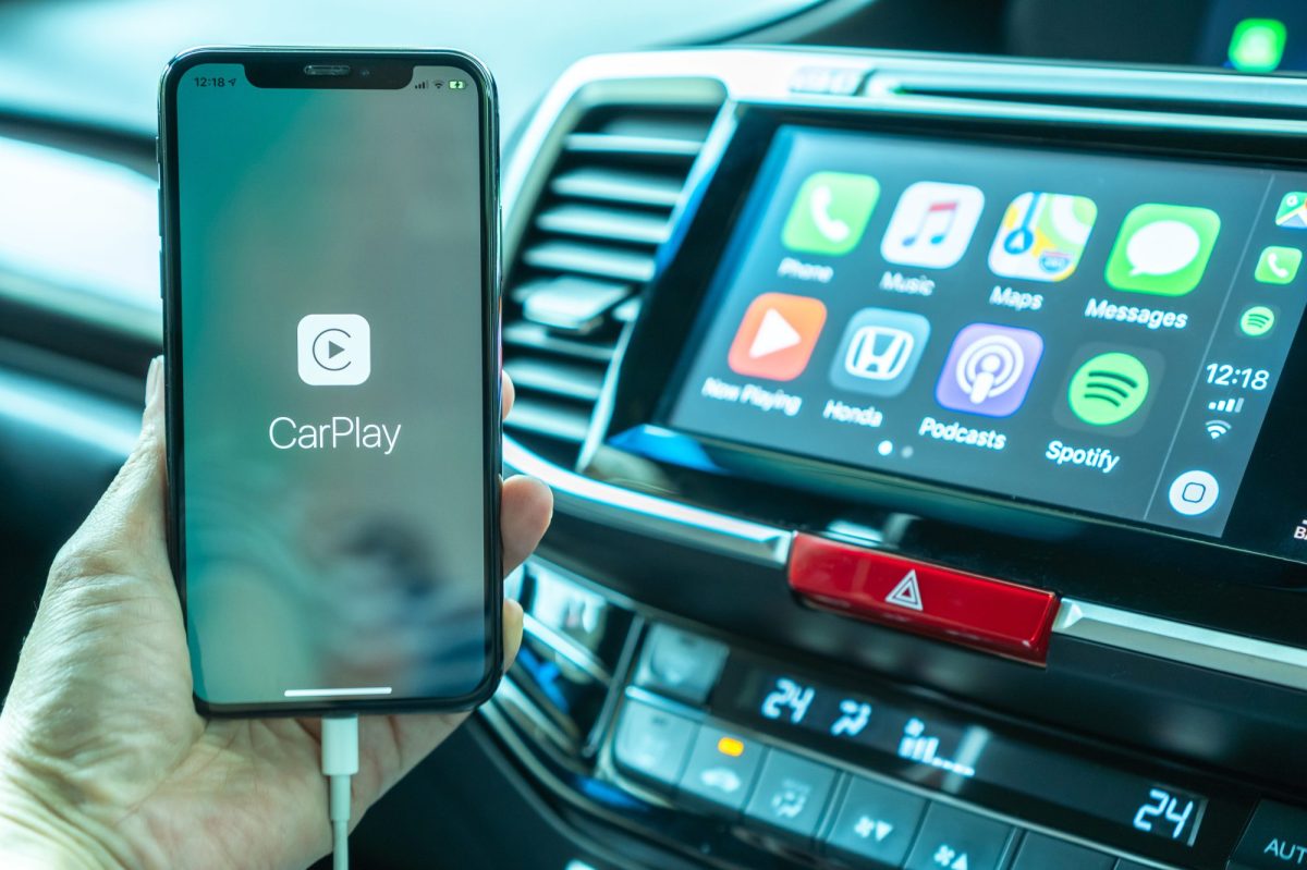 Apple CarPlay wird auf einem Handy angezeigt. Im Hintergrund ist ein Auto-Display mit verschiedenen Apps zu sehen.